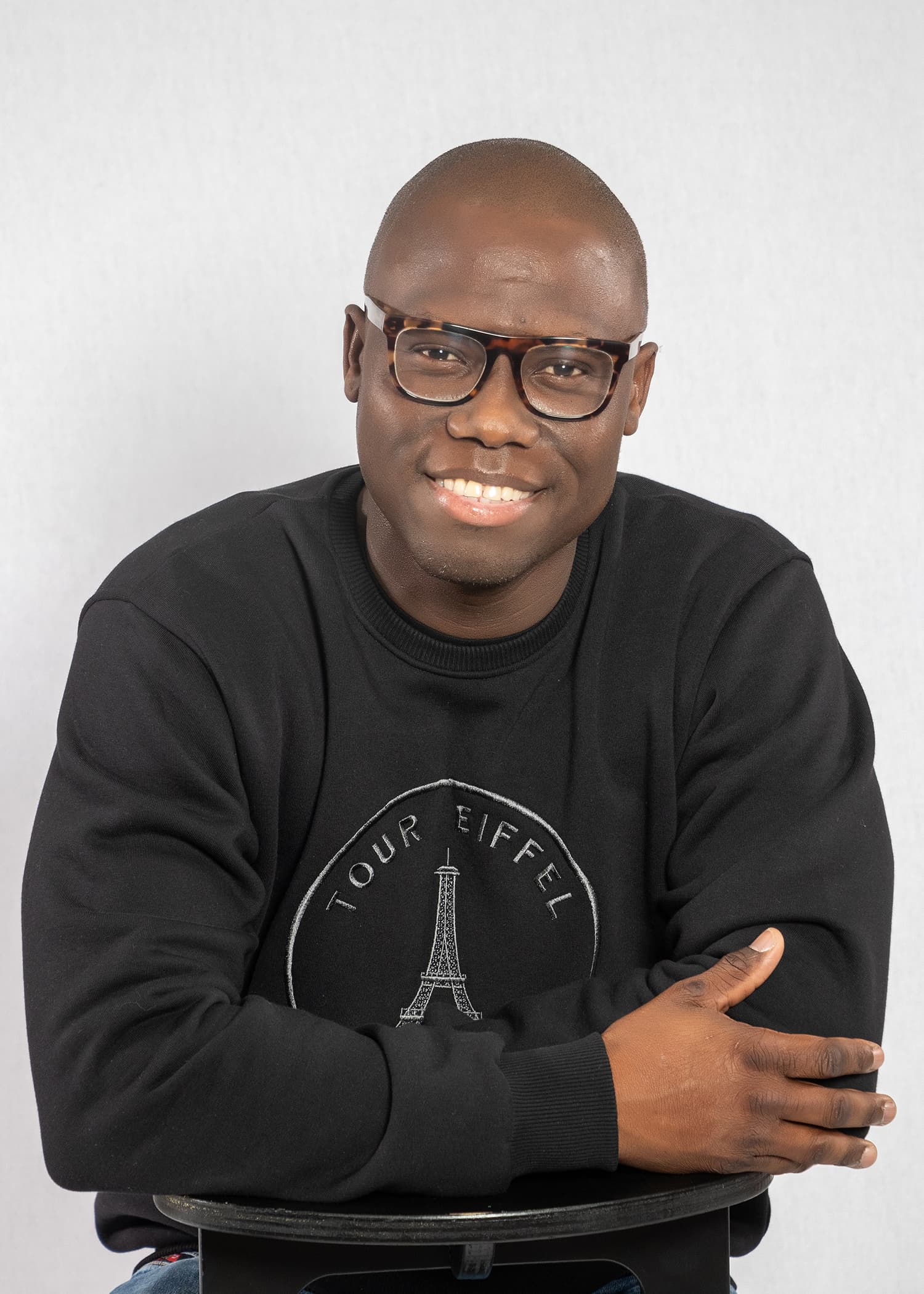 Smiling Black man with tortoiseshell glasses wearing a black sweatshirt with an Eiffel Tower graphic.